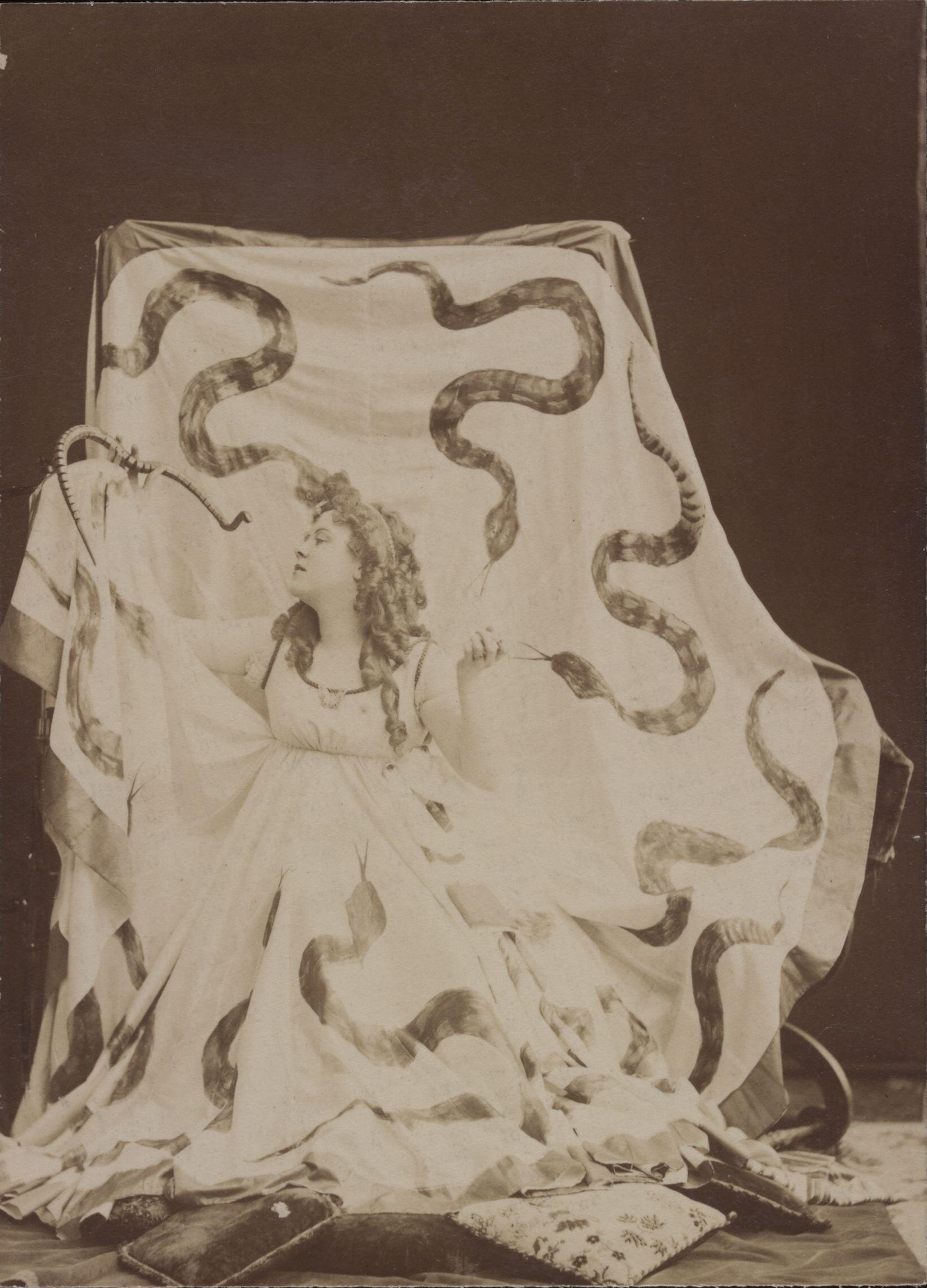 A woman in a long, flowing dress covered with painted snakes. She is holding a wooden snake in one hand, and lookint into its face. She has long, curled hare, and there are pillows spread out on the floor around her.