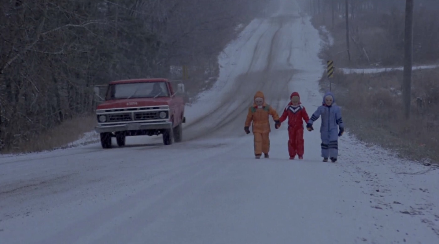 Three children walking hand in hand down the side of a street in the snow. A truck is driving by.